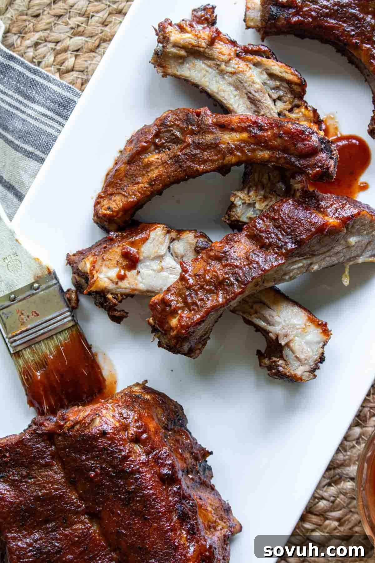 A white plate with saucy, Slow Cooker Ribs, some separated and others in a rack, next to a sauce brush with barbecue sauce.