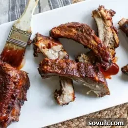 A plate of Slow Cooker Ribs with a basting brush, some ribs whole and others cut individually, set on a white rectangular plate with a cloth napkin partially visible in the background.