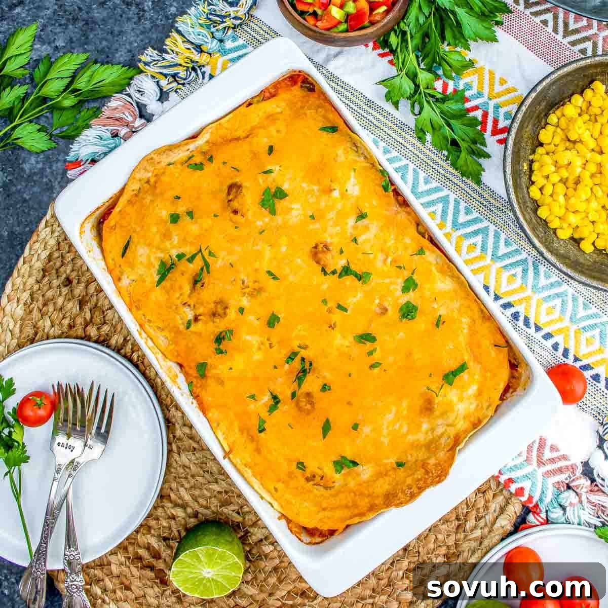 Overhead view of a homemade Chicken Tortilla Casserole in a white dish surrounded by fresh herbs, a bowl of corn, and salad, served on a colorful tablecloth.