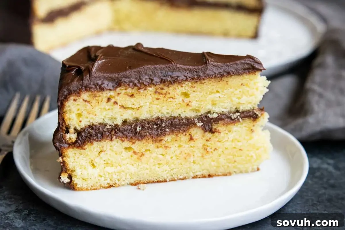 A slice of yellow layer cake with rich chocolate frosting on a white plate, with the rest of the beautifully frosted cake in the background, ready to be served.