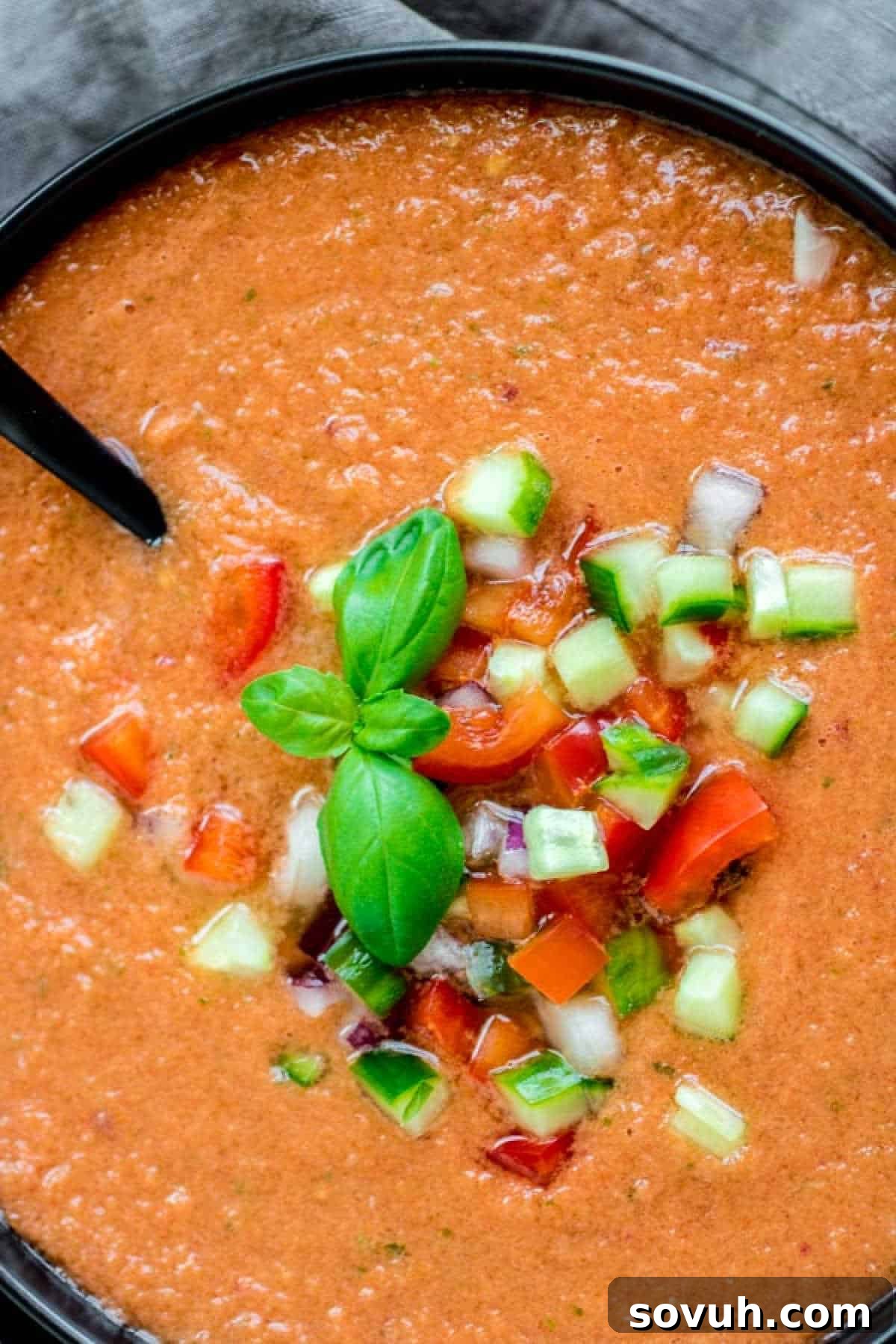 A black ceramic bowl filled with vibrant red Gazpacho Soup, with a spoon resting in it, ready for enjoyment.