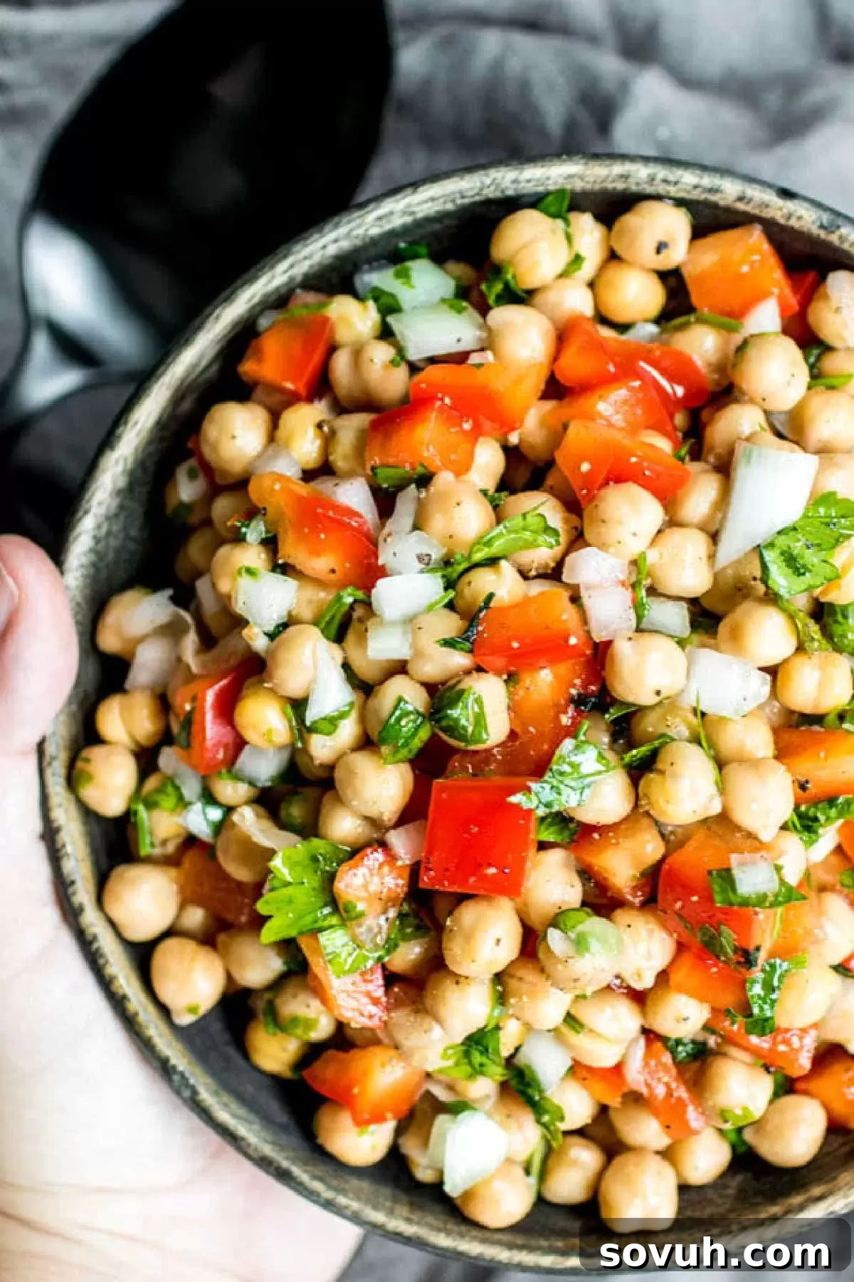 A hand holding a rustic wooden bowl filled with freshly made Mediterranean Chickpea Salad, showcasing its colorful ingredients and ready for a meal