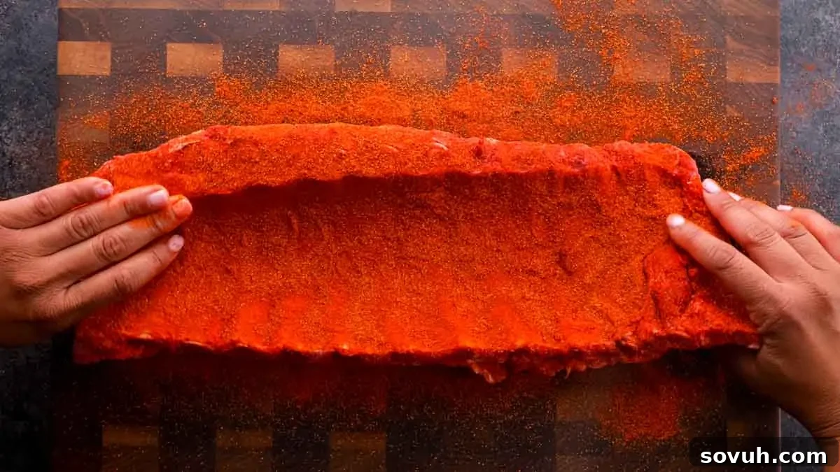 The Ultimate Memphis Dry Rub 4 Hands coating a rack of St Louis-Style Ribs with a vibrant red spice rub on a rustic wooden cutting board, preparing them for the grill.