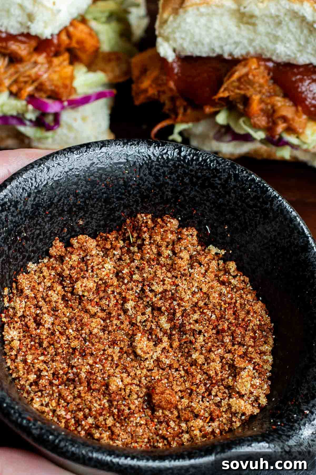 The Ultimate Memphis Dry Rub 5 A close-up of a black bowl filled with a Memphis Dry Rub. In the background, there are delicious-looking sandwiches with shredded meat and fresh greens, suggesting meal ideas.