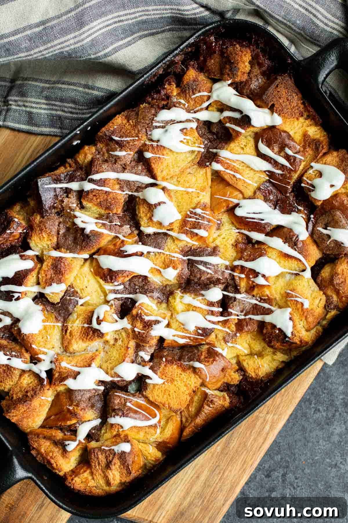 A whole Cinnamon Roll French Toast Casserole in a rustic cast iron pan, beautifully baked to a golden hue and generously covered with a smooth, white cream cheese icing, ready to be served.