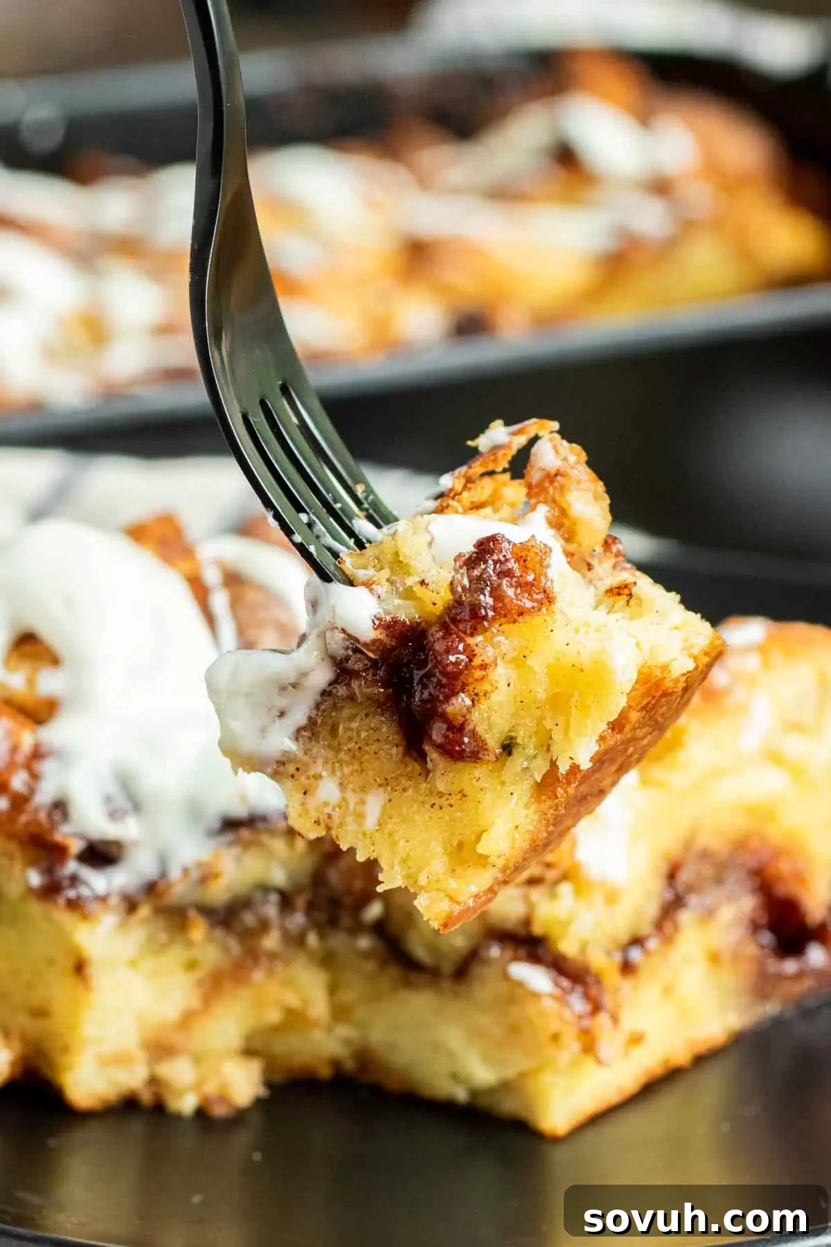 A fork piercing a piece of gooey, cinnamon-laden Cinnamon Roll French Toast Casserole, with visible cream cheese icing, ready to be lifted and savored, emphasizing its rich texture.