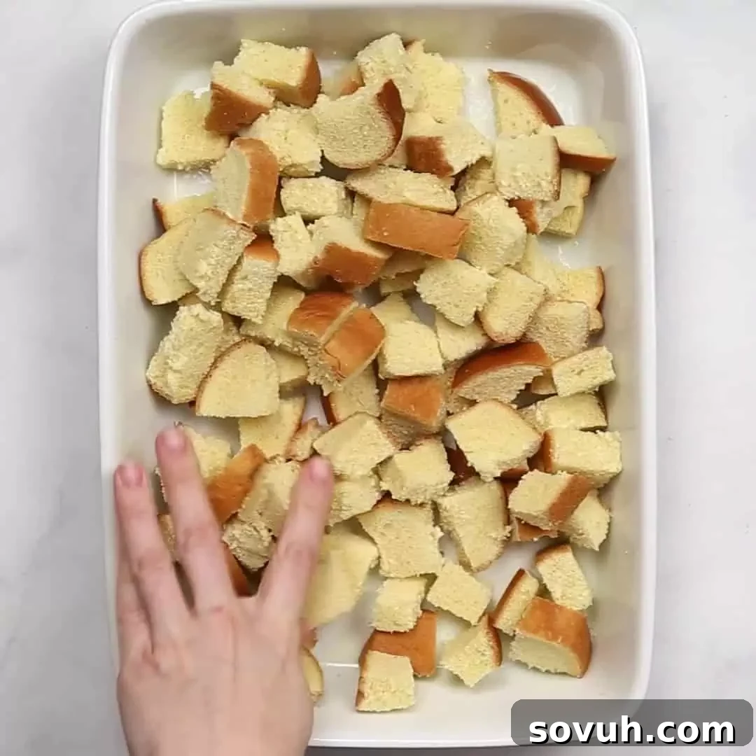 A hand gently tearing brioche bread into large, even chunks inside a baking dish, preparing it for the Cinnamon Roll French Toast Casserole.
