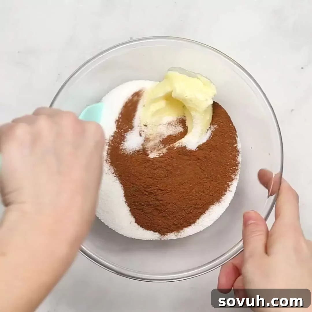 A person's hands skillfully mixing softened butter, granulated sugar, and ground cinnamon in a clear glass bowl to create the rich, aromatic cinnamon-sugar filling.