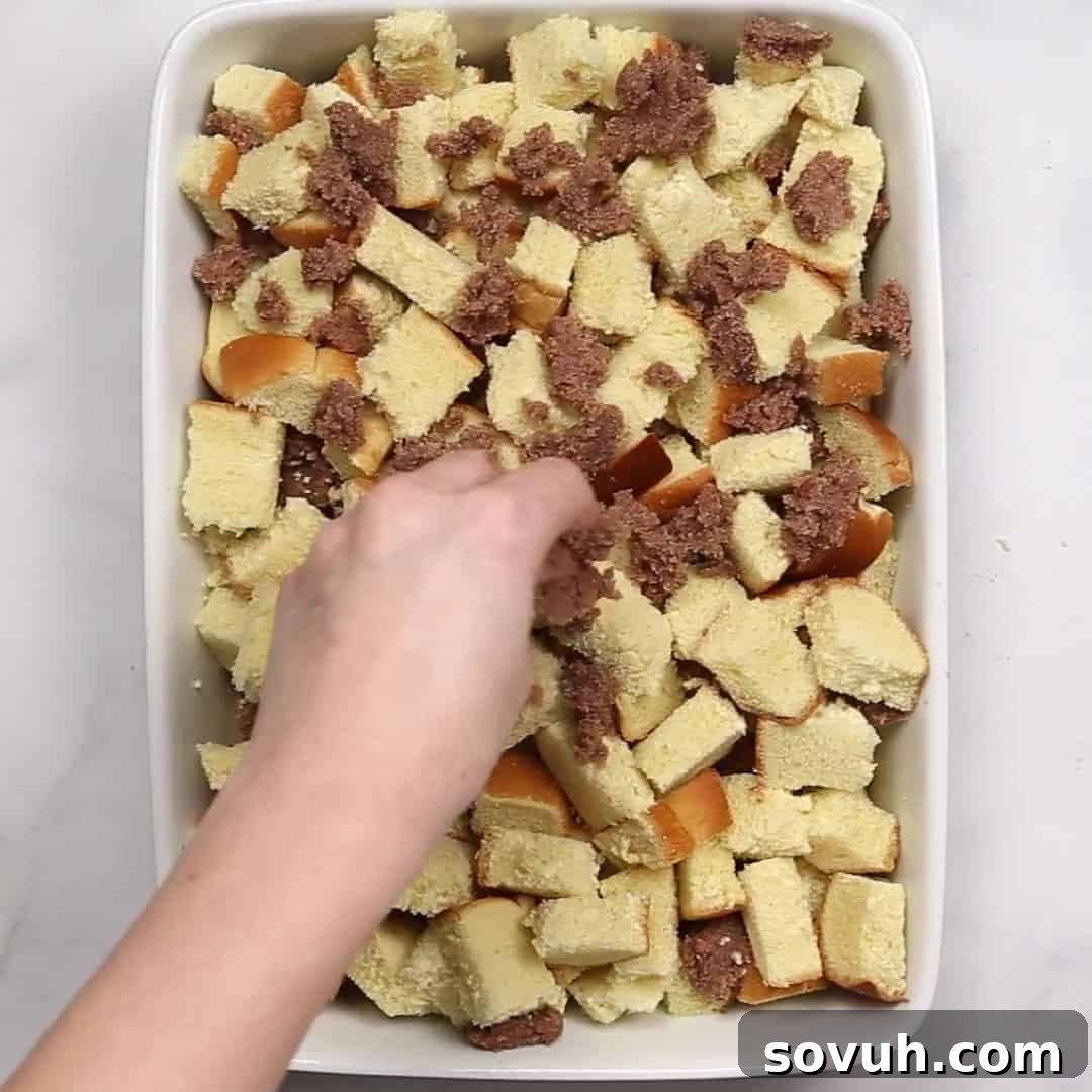 A baking dish being filled with layers of brioche bread chunks, dotted generously with spoonfuls of the cinnamon-sugar butter mixture, showing the assembly process.