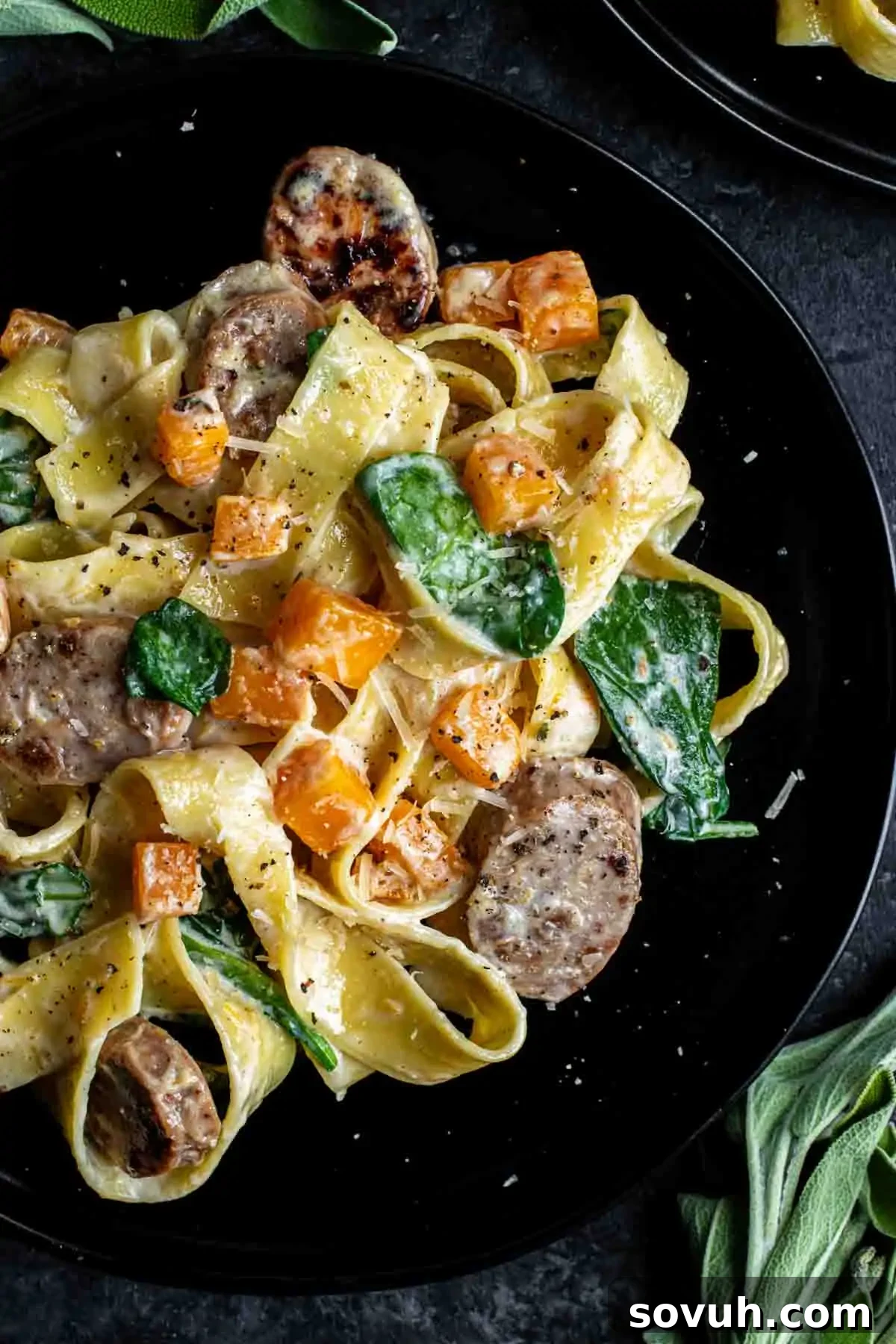 Delicious Creamy Butternut Squash and Sausage Pasta A plate of tagliatelle pasta with sliced sausage, cubes of butternut squash, spinach leaves, and a creamy sauce, garnished with black pepper.