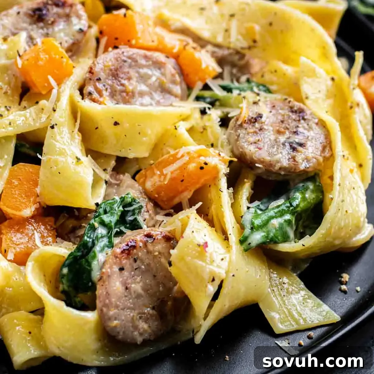 Creamy Butternut Squash and Sausage Pasta Ready to Eat Close-up of a plate with pappardelle pasta, sliced sausage, spinach, and chunks of orange squash, garnished with grated cheese and black pepper.