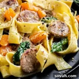 Close-up of a plate with pappardelle pasta, sliced sausage, spinach, and chunks of orange squash, garnished with grated cheese and black pepper.