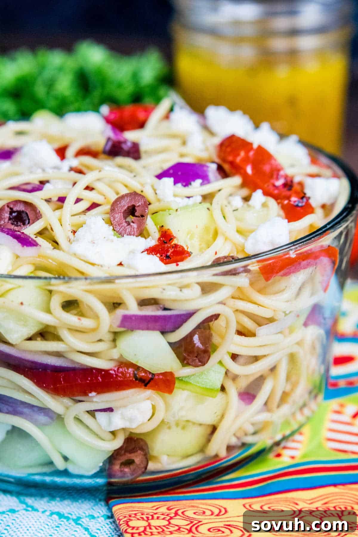 glass bowl with Greek Spaghetti Salad with jar of dressing behind bowl