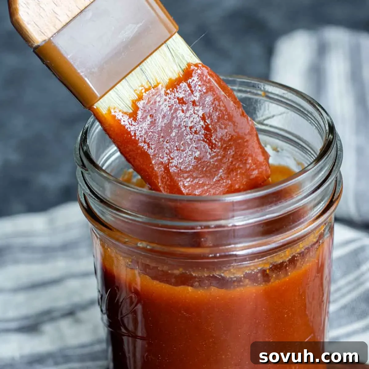 A brush applying rich, reddish-brown Memphis BBQ Sauce from a jar onto a textured dark background, highlighting its thick, glossy consistency.