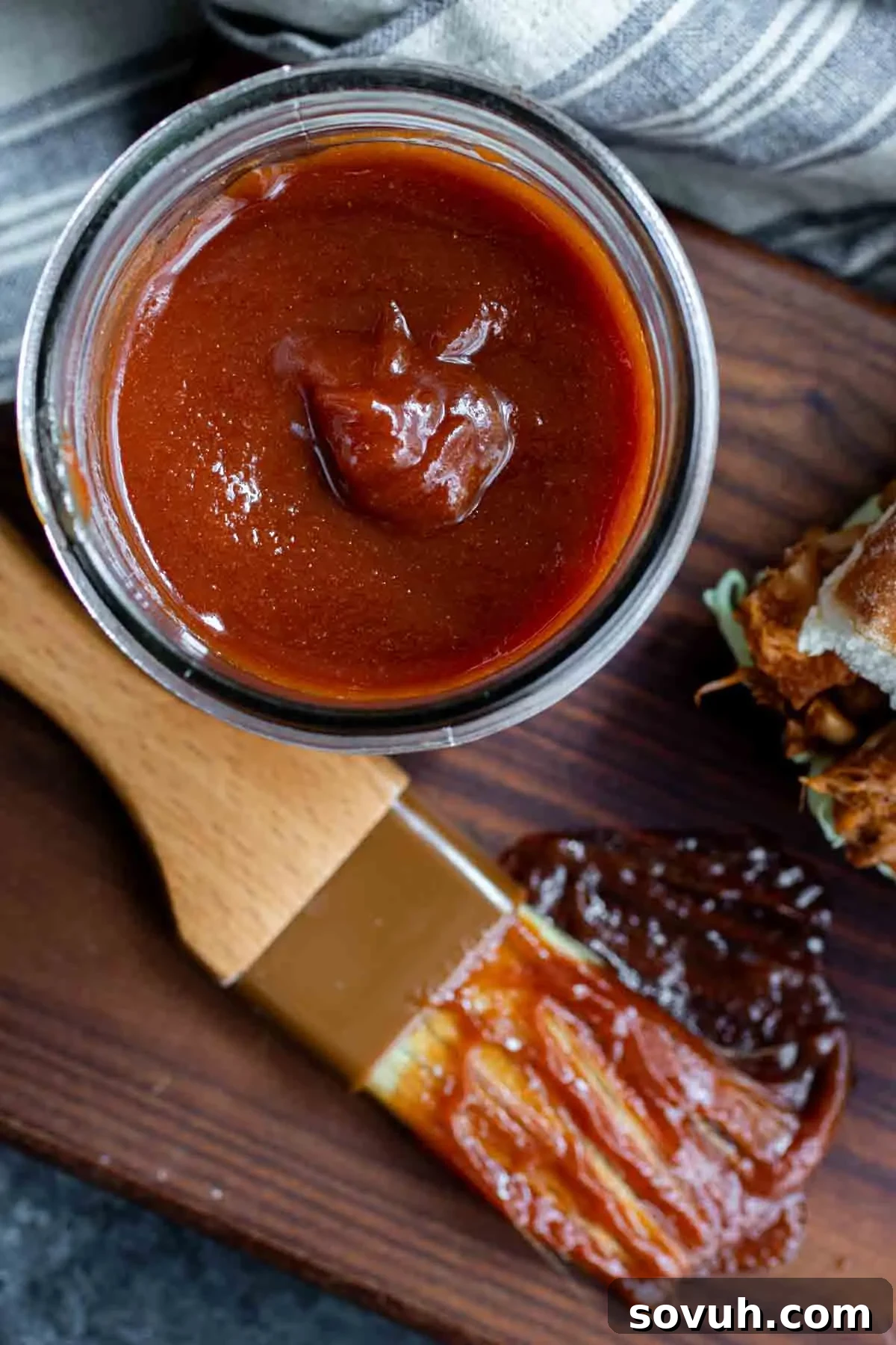 Top-down view of a jar filled with glistening Memphis BBQ Sauce, with a spoon resting in it, next to a basting brush and a small amount of sauce smeared artistically on a wooden surface.