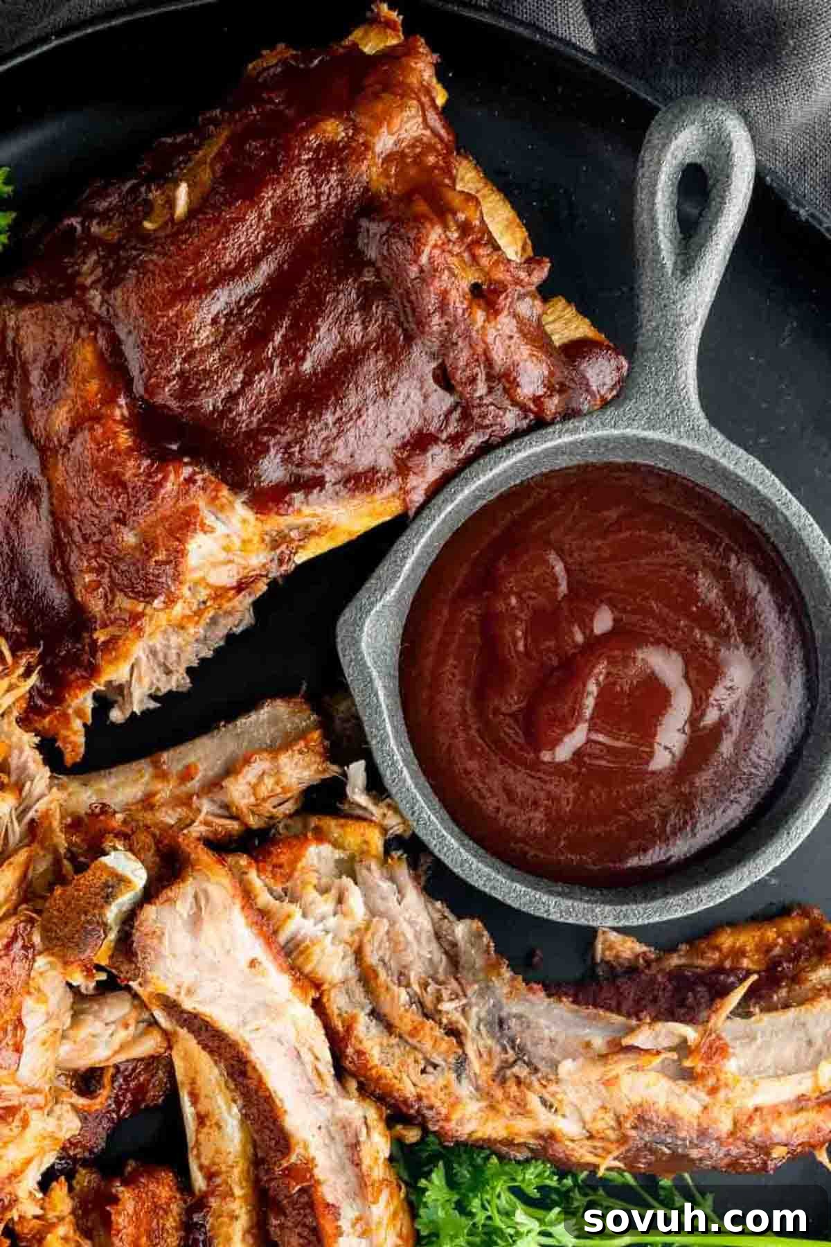 A delicious plate of barbecue ribs, glistening with Memphis BBQ Sauce, served with a small cast iron skillet of extra sauce on a dark background, viewed from above.