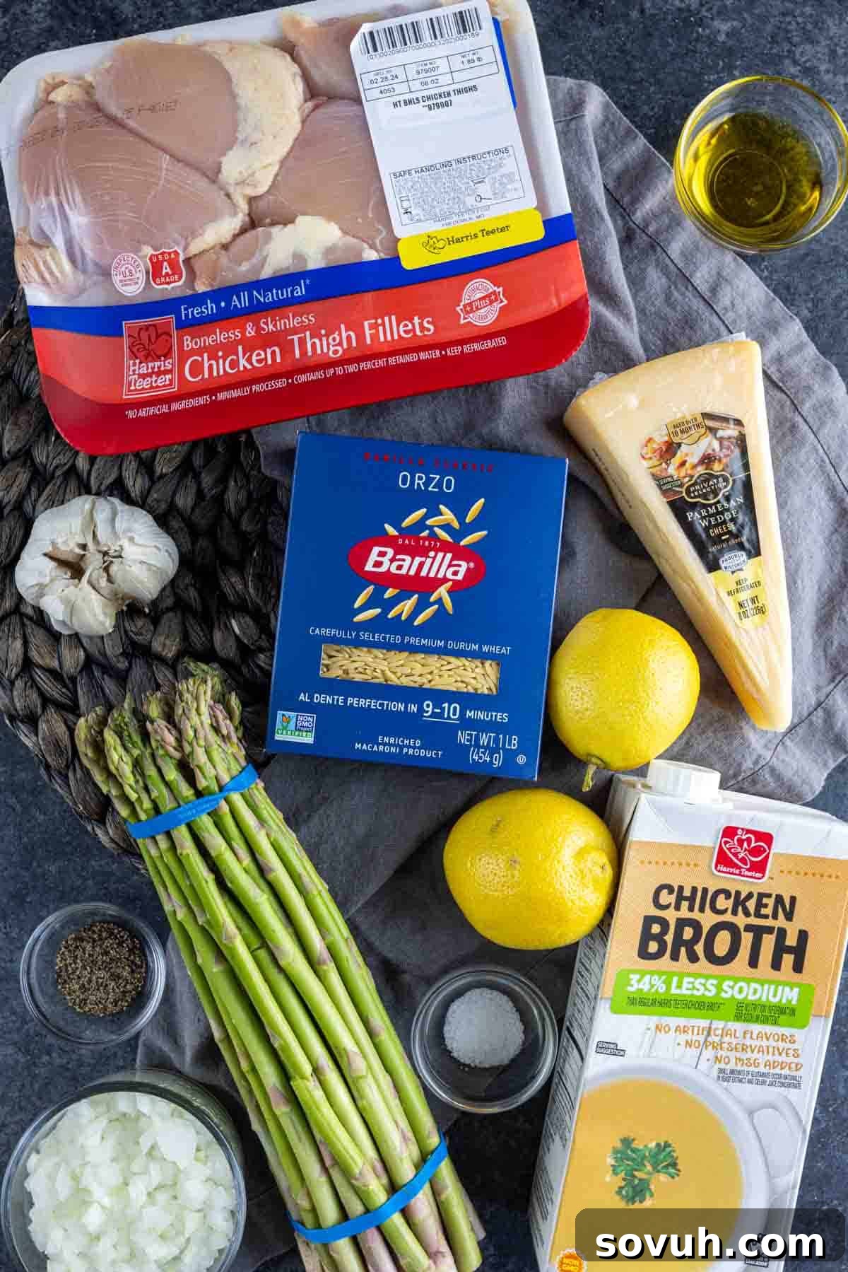 Parmesan Chicken Orzo Ingredients for a recipe including chicken thigh fillets, orzo pasta, parmesan cheese, lemons, asparagus, garlic, chicken broth, olive oil, and spices on a dark surface, laid out for cooking.