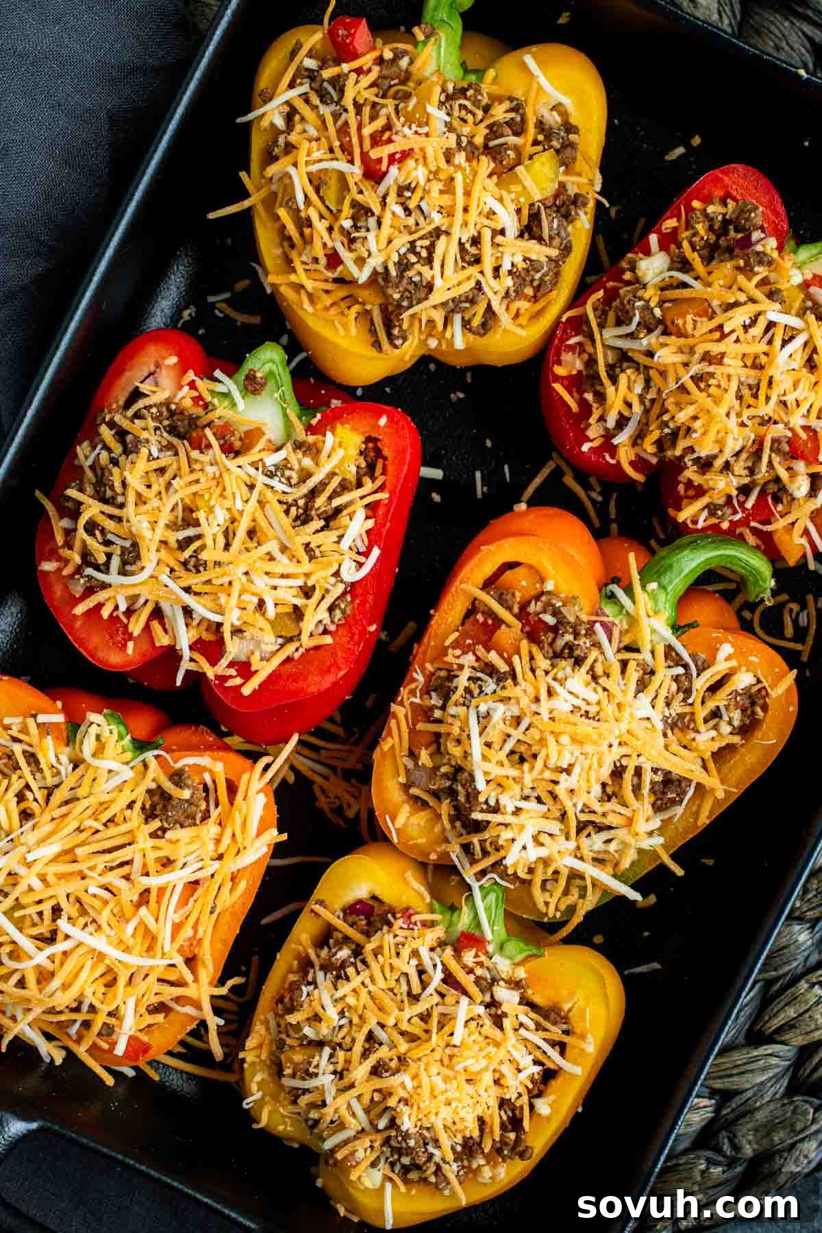 Six colorful bell pepper halves in a baking tray, stuffed with ground meat and topped with shredded cheese, ready for baking.