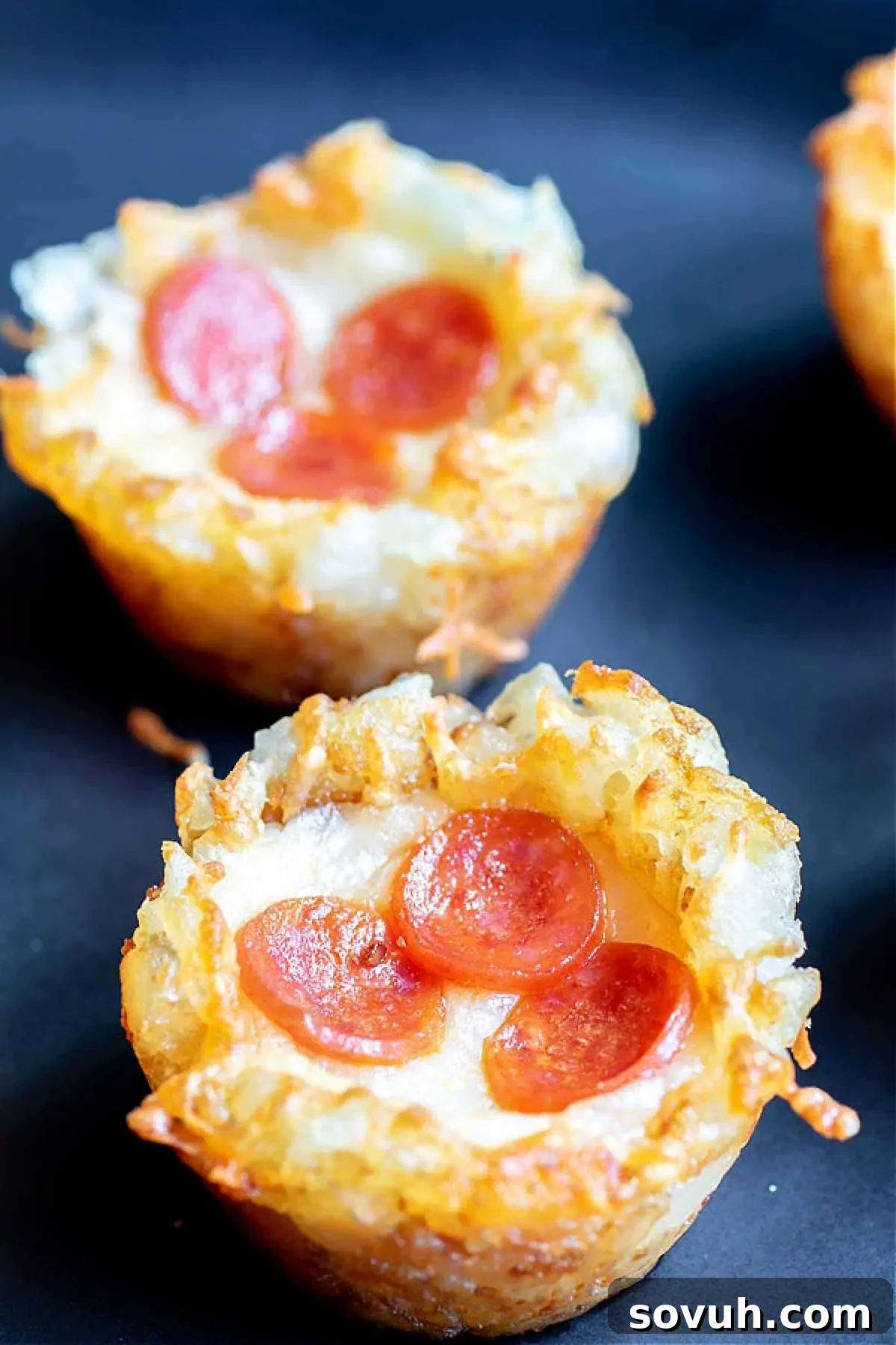 Three pepperoni pizza muffins on a dark surface, showing melted cheese and pepperoni slices on top of a crispy crust.