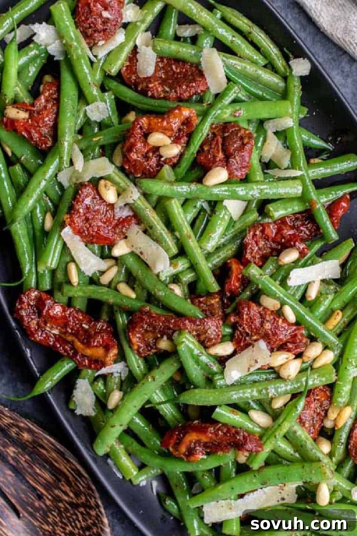 A dish of green beans with sun-dried tomatoes, pine nuts, and shaved cheese on a black plate.