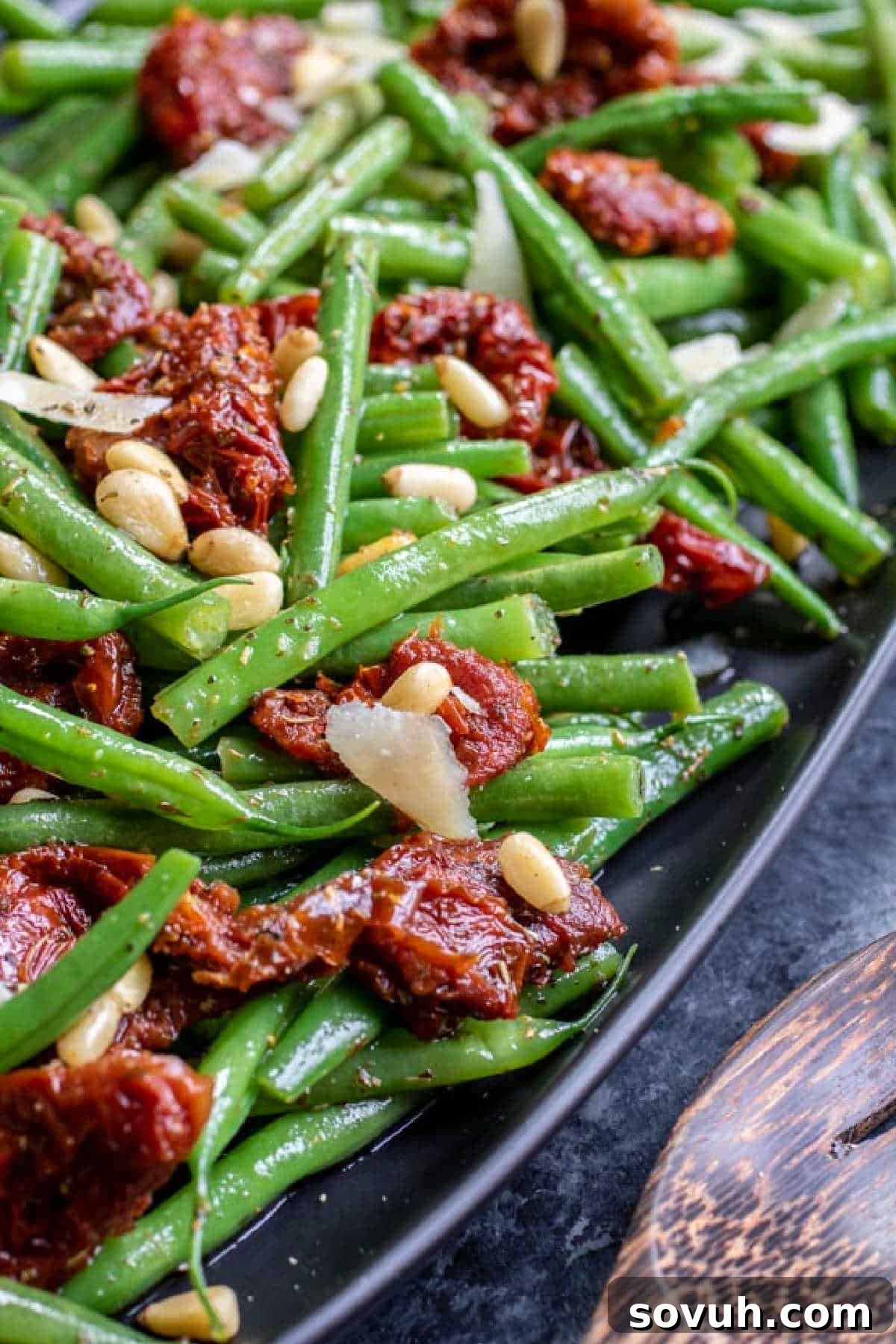 A close-up of a dish with sautéed green beans, sun-dried tomatoes, pine nuts, and sliced onions on a dark plate.