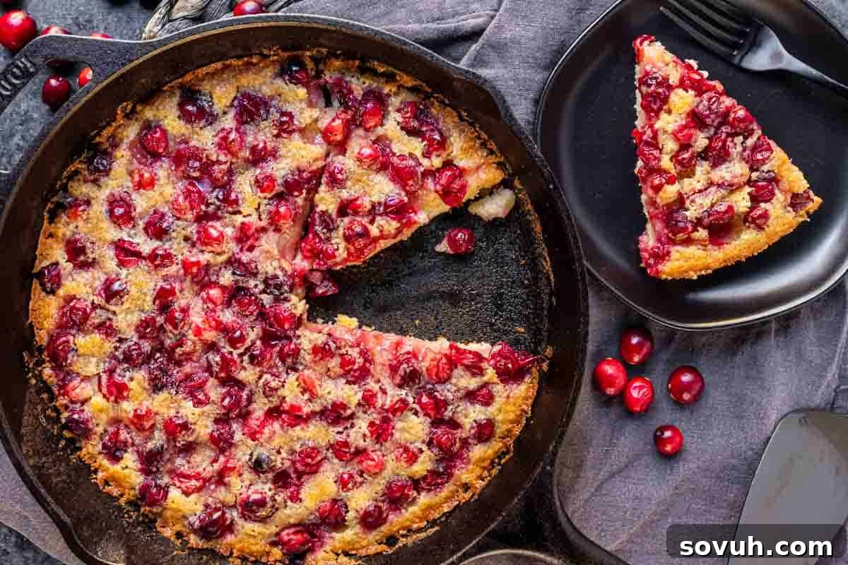 A beautifully baked cranberry buckle in a cast iron skillet, with a slice removed and placed on a black plate, garnished with scattered fresh cranberries and a sprig of rosemary.