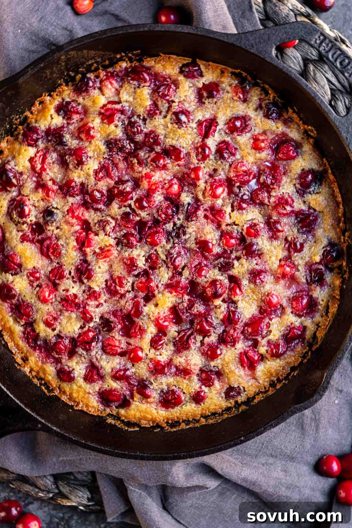 A baked Cranberry Buckle in a cast iron skillet, set on a dark woven mat, showcasing its golden-brown crust and glistening cranberries.