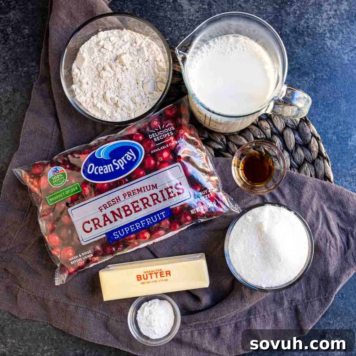 Key ingredients for a Cranberry Buckle laid out on a table: all-purpose flour, whole milk, fresh red cranberries, vanilla extract, granulated sugar, and a stick of butter.