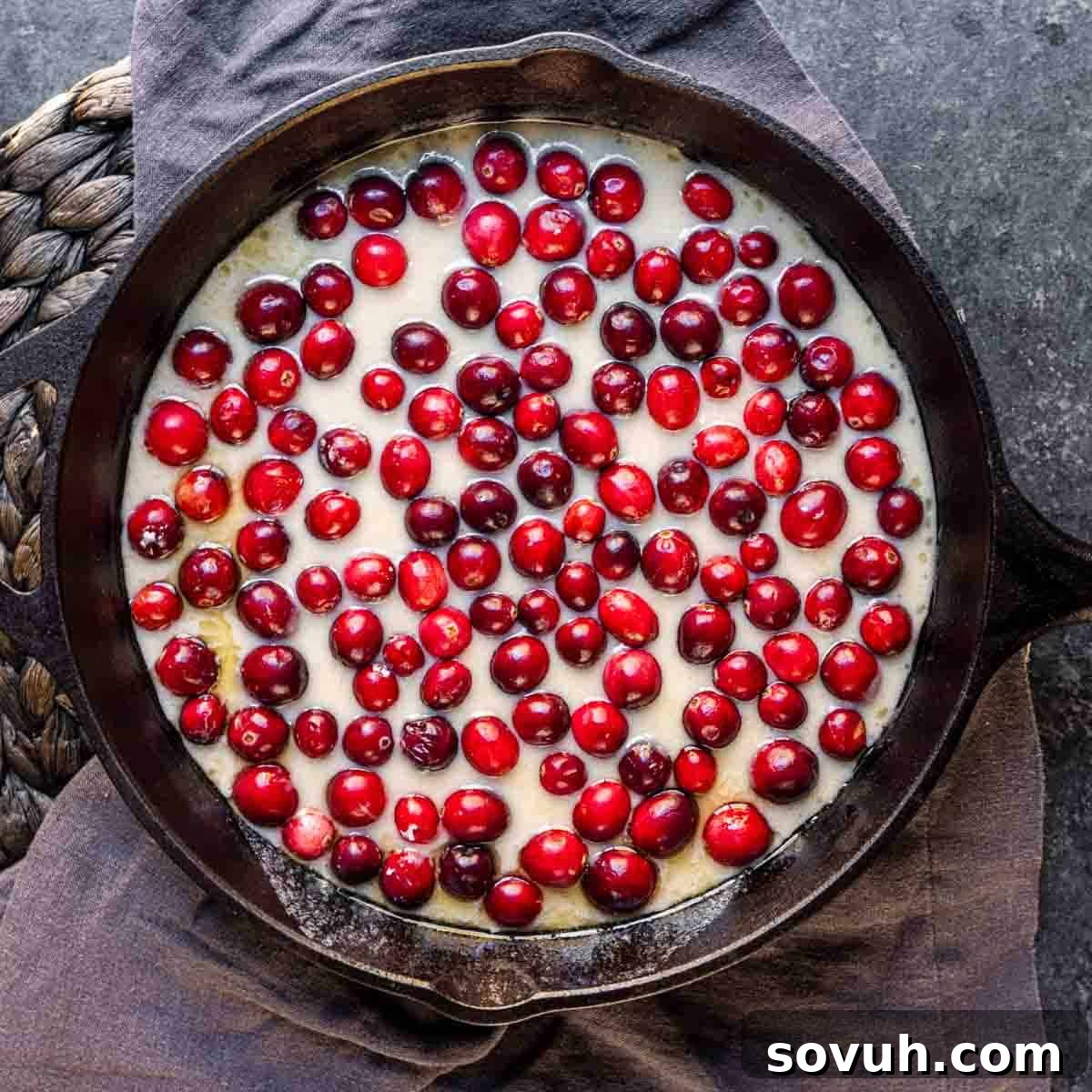 A cast iron skillet filled with smooth cake batter, generously topped with a layer of fresh, bright red cranberries, ready for baking.