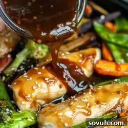 Sauce being poured over cooked chicken, surrounded by vegetables like broccoli, carrots, and snap peas, garnished with sesame seeds.