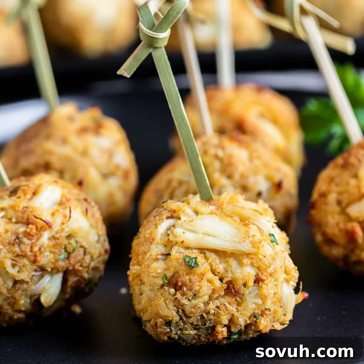 Close-up of golden-brown crab cakes on skewers, placed on a black plate, garnished with a sprig of parsley in the background.