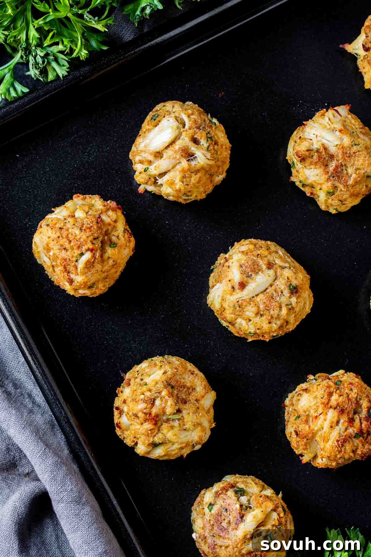 Baked crab cakes on a black baking sheet surrounded by parsley.