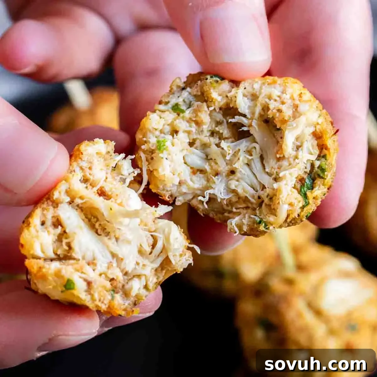Hands holding a broken crab cake, revealing flaky crab meat inside.
