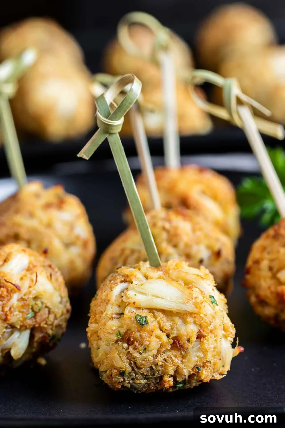 Close-up of crab cakes on a black plate, each pierced with a decorative wooden skewer.
