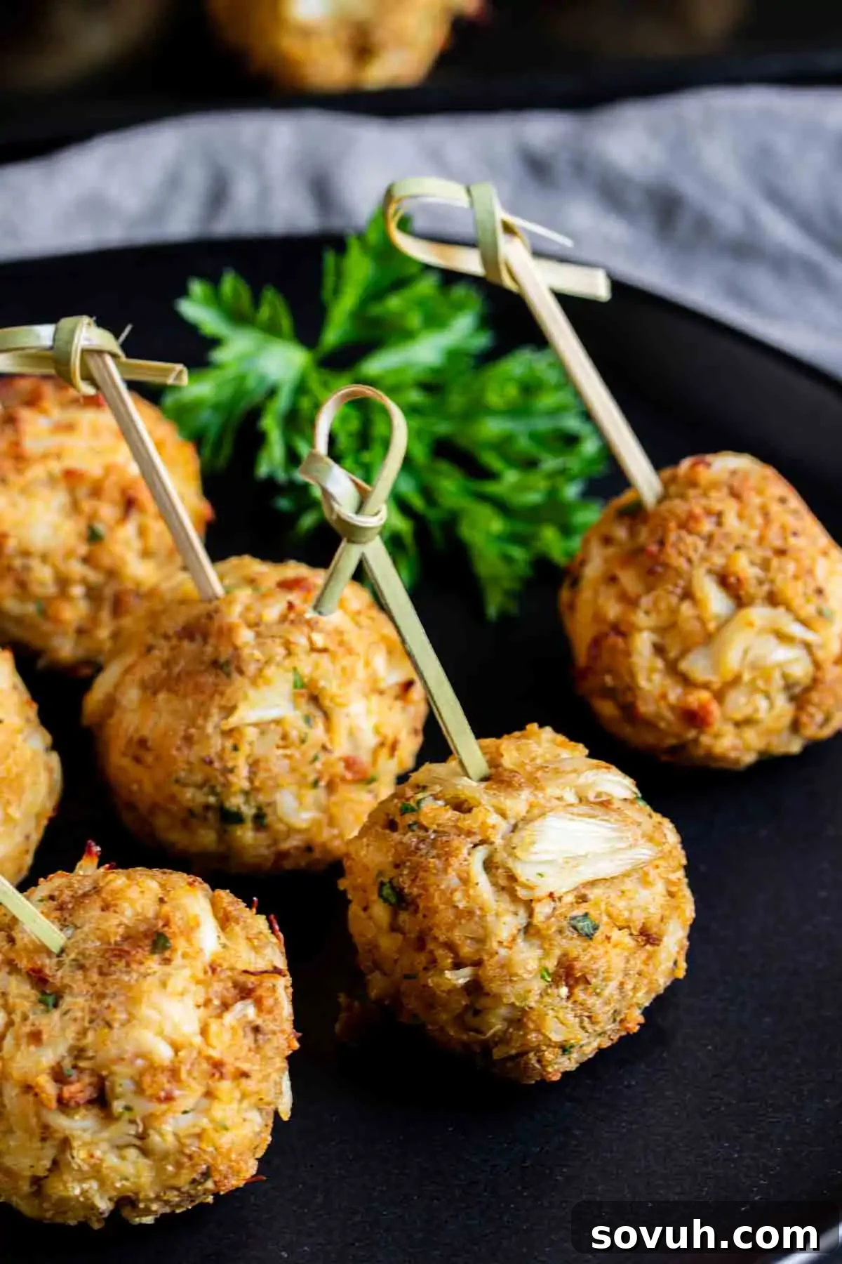 Close-up of five crab cakes on a black plate, each skewered with a bamboo pick. A parsley garnish is in the background.