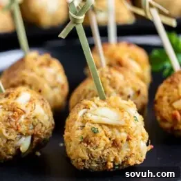 Close-up of golden-brown crab cakes on skewers, placed on a black plate, garnished with a sprig of parsley in the background.