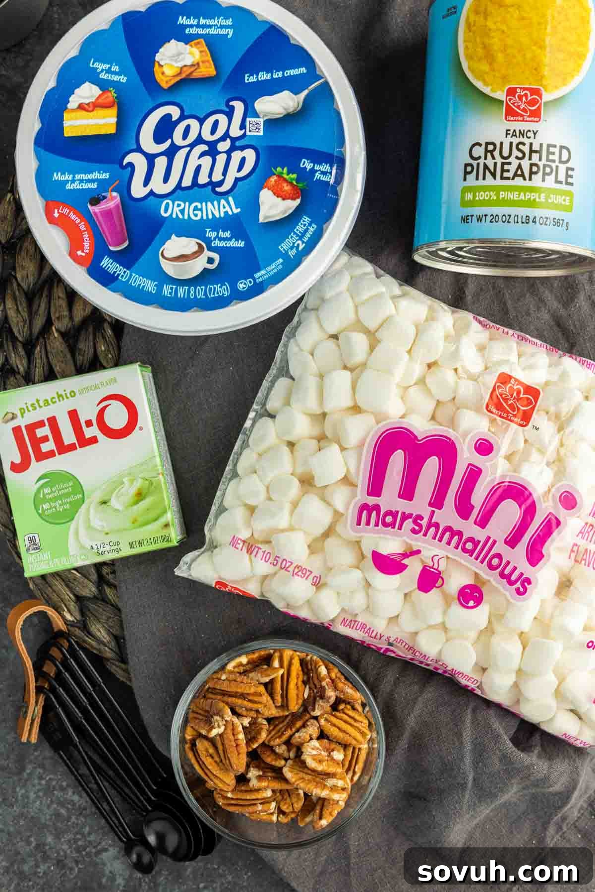 A flat lay of all the essential ingredients for Watergate Salad: a box of pistachio pudding, a can of crushed pineapple, a tub of Cool Whip, a bag of marshmallows, and chopped pecans.