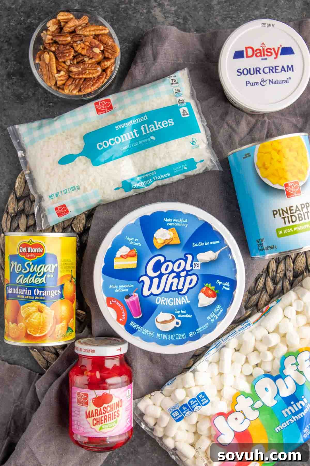 A collection of fresh and canned ingredients for Ambrosia Salad, neatly arranged on a counter, including Cool Whip, sour cream, various fruits, marshmallows, and coconut.