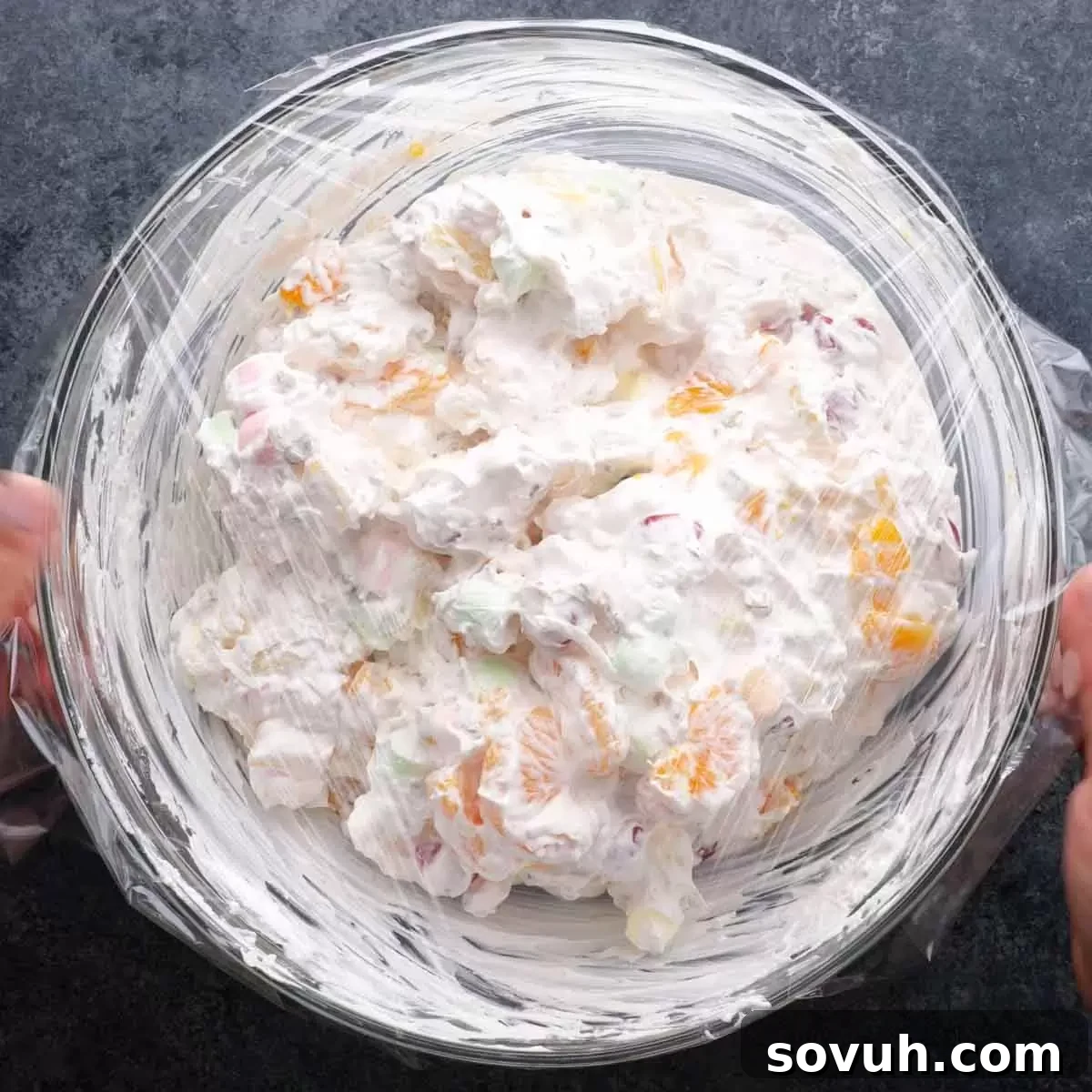 Step-by-step image of the Ambrosia Salad in a bowl being covered with plastic wrap, ready for chilling in the refrigerator.