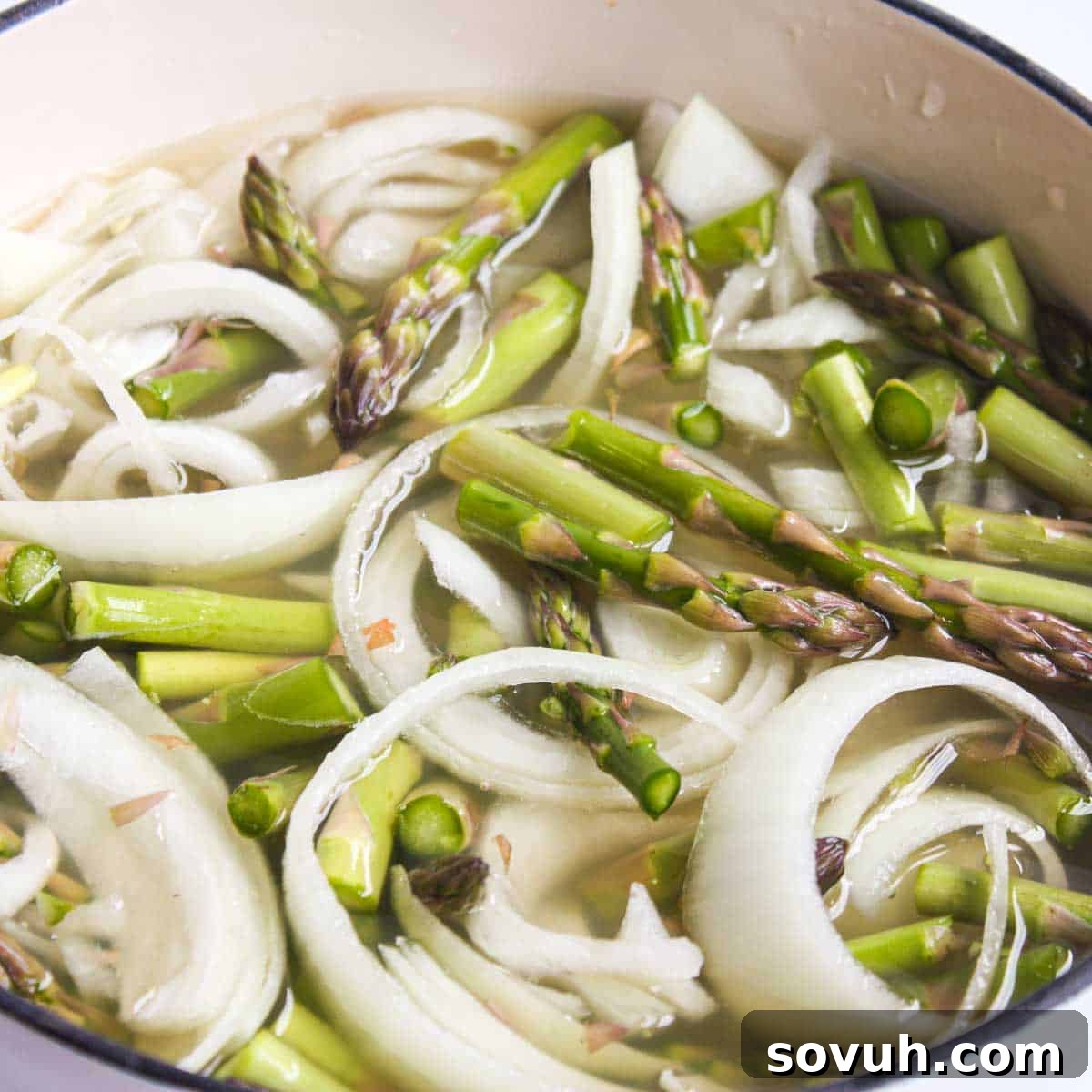 Fresh ingredients for Creamy Asparagus Soup, including asparagus, onion, garlic, butter, and broth, in a large pot, ready for cooking.