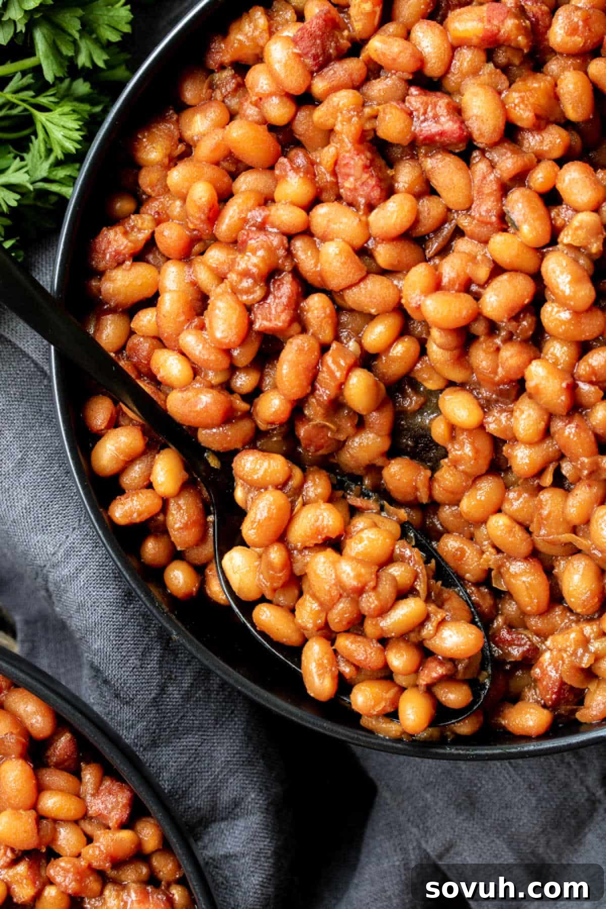 A black spoon rests invitingly in a rustic bowl of finished Instant Pot Boston baked beans, garnished with crispy pieces of bacon and fresh green parsley leaves, suggesting a delicious and hearty meal.