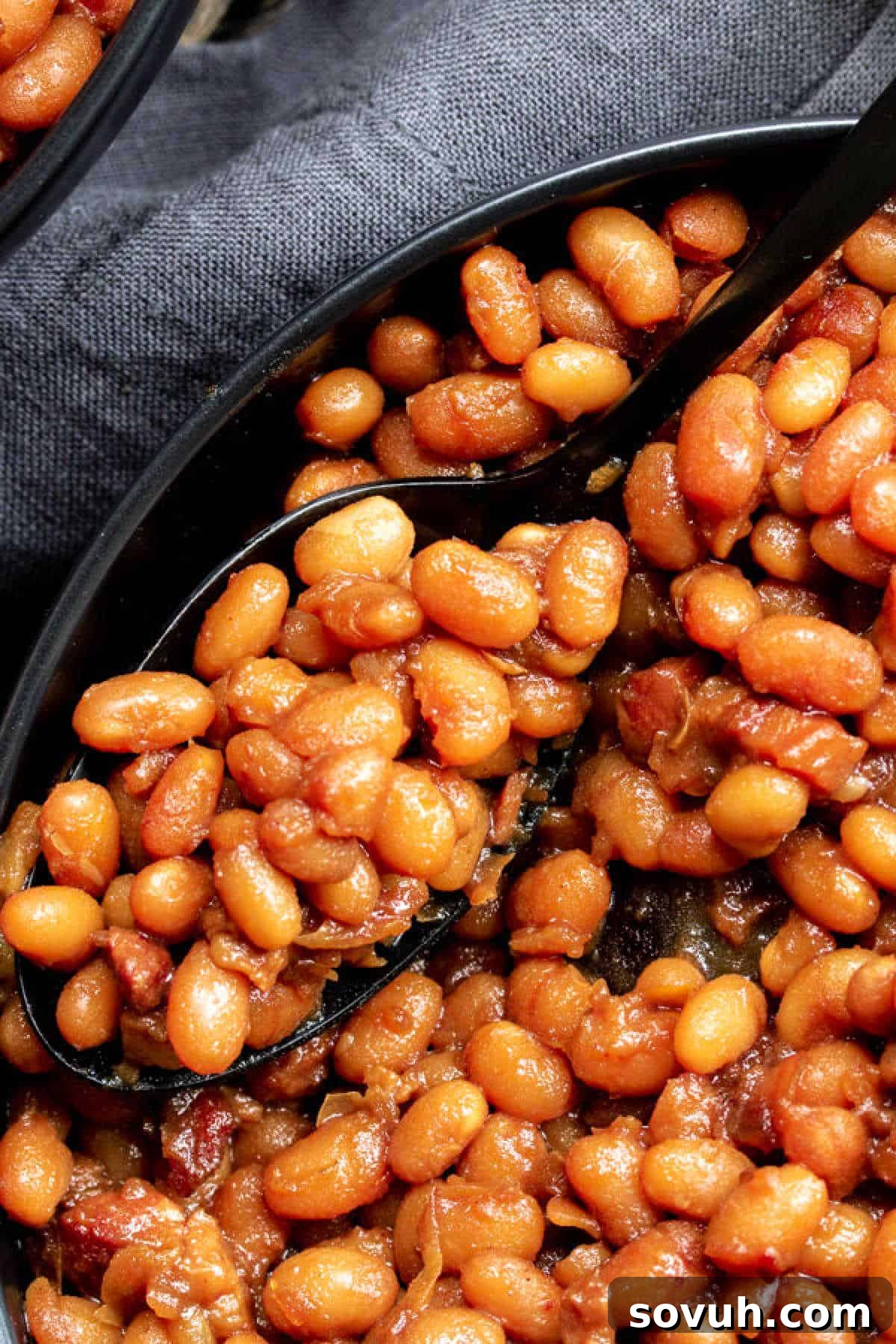 Close-up of a rustic bowl filled with perfectly cooked Instant Pot Boston baked beans, glistening with a rich, dark sauce. A black spoon is scooping out a generous portion, showcasing the even coating and tender texture of the beans.