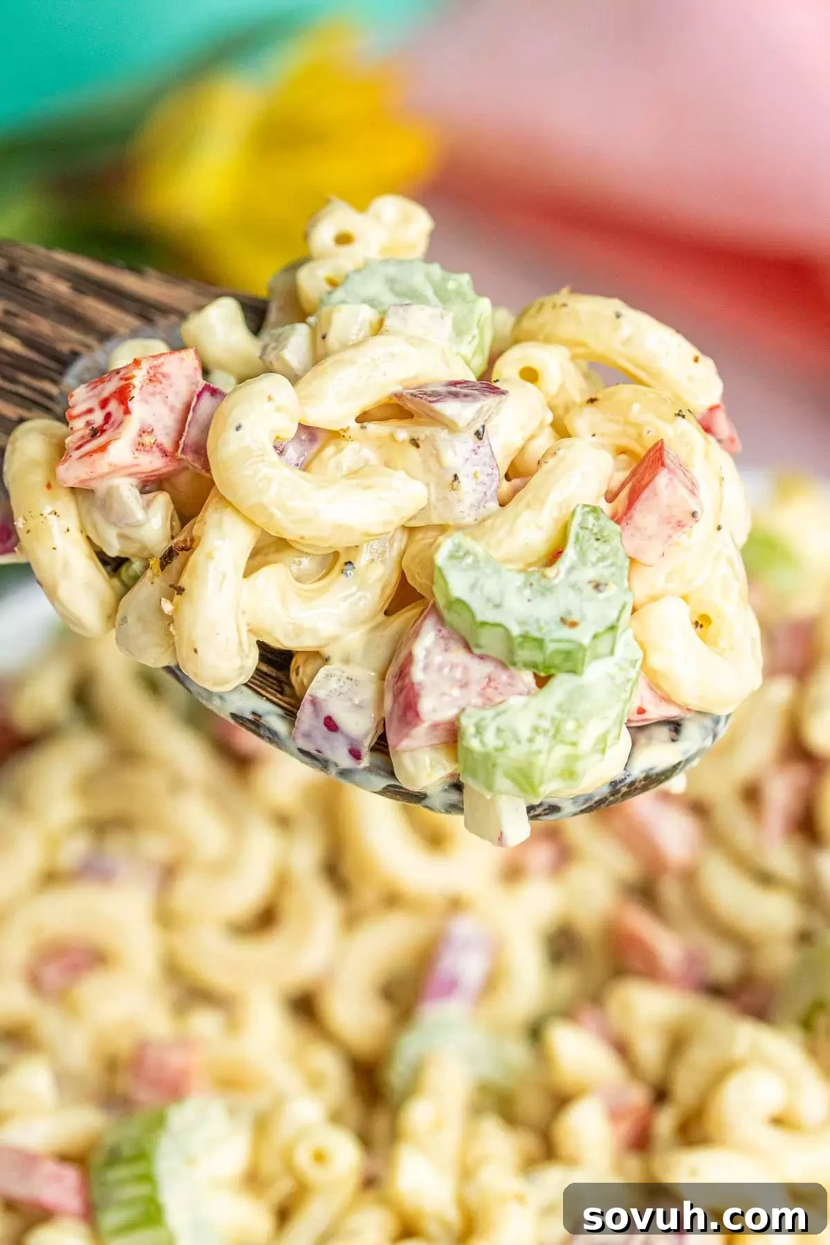 A wooden spoon scooping a generous portion of classic macaroni salad from a large bowl, highlighting its creamy texture and visible vegetable pieces.