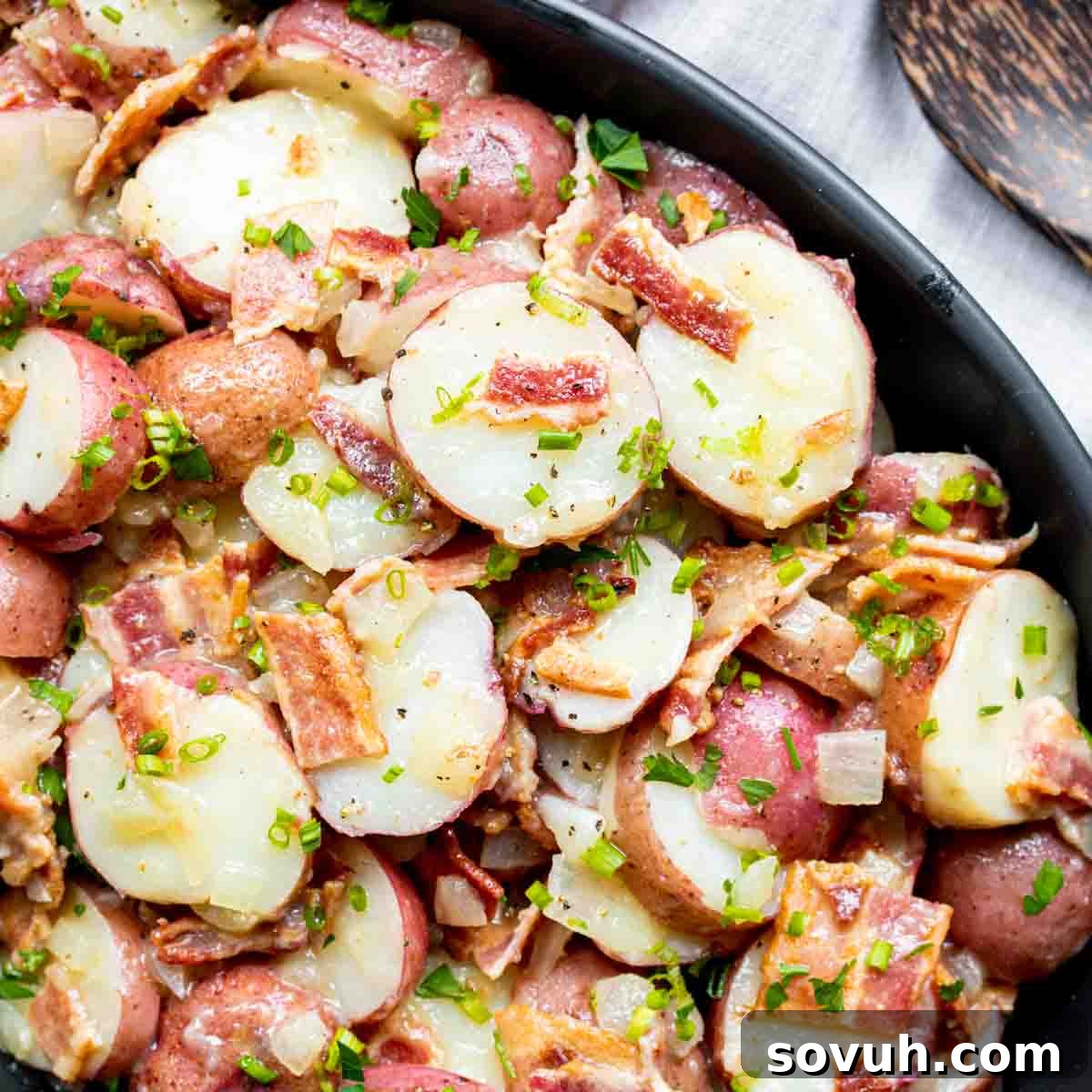 A rustic black bowl filled with warm German Potato Salad, garnished with fresh chives and crispy bacon bits, ready to be served.