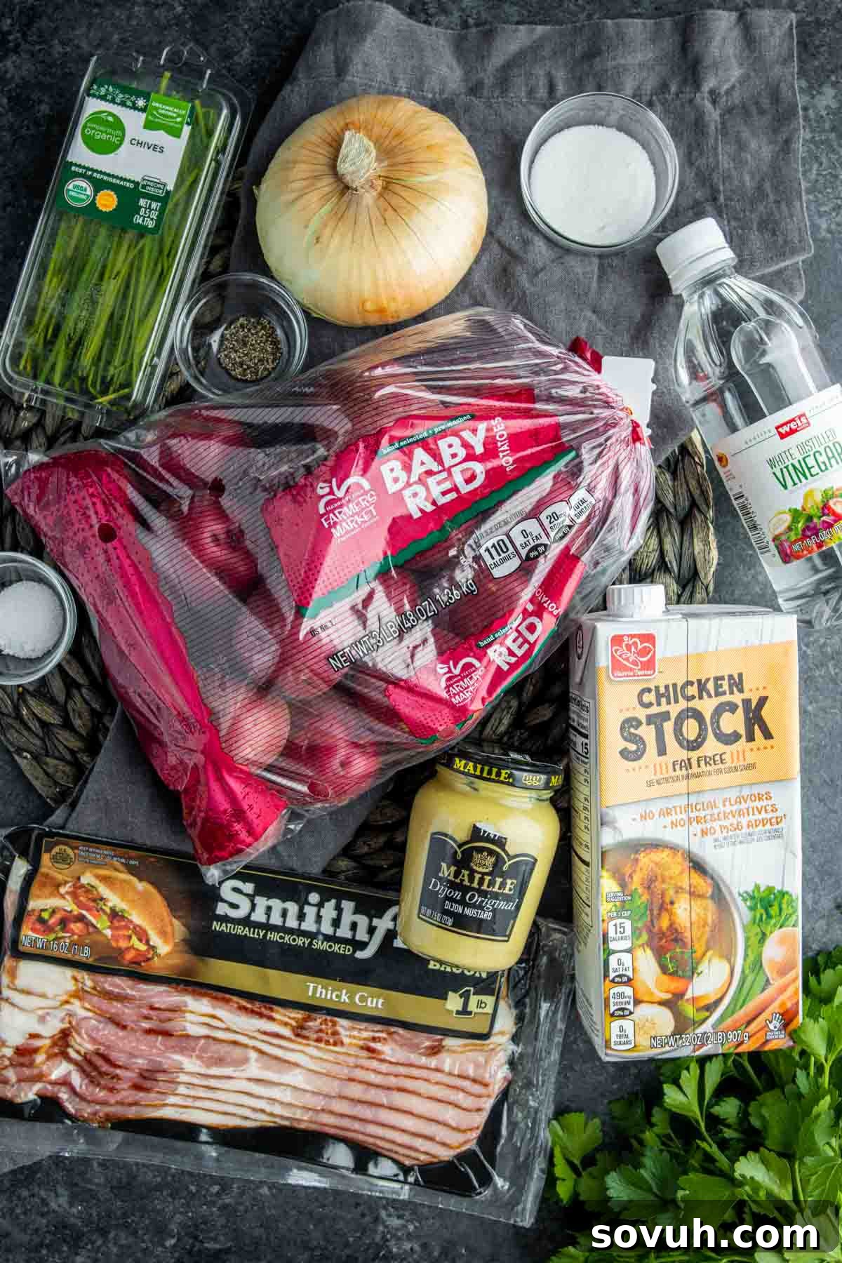 Close-up of all the fresh ingredients laid out on a wooden board, including red potatoes, fresh herbs, bacon, onions, and various liquids, ready for making warm potato salad.