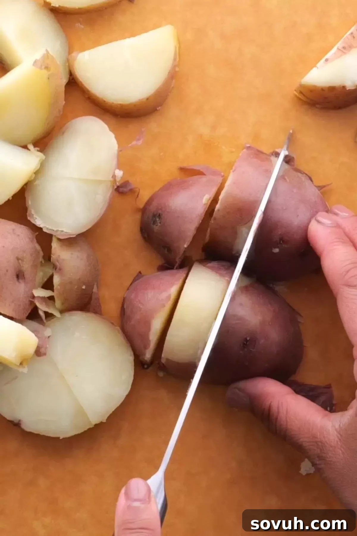 Close-up of freshly boiled red potatoes being carefully sliced into half-moon shapes on a cutting board, a key step for making German potato salad.