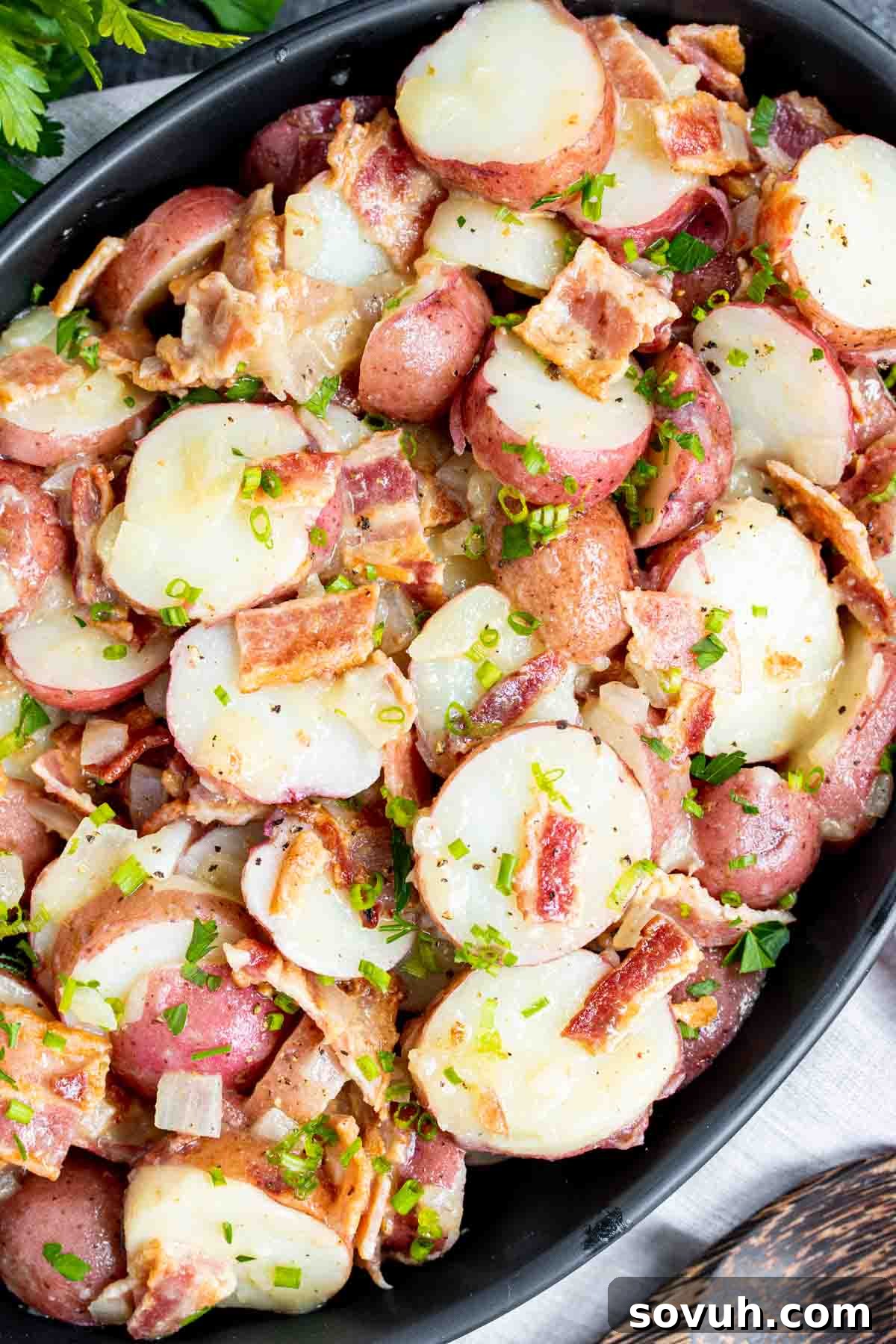 Close-up of a serving of German potato salad in a black dish, garnished with fresh herbs and visible bacon bits, ready for a meal.
