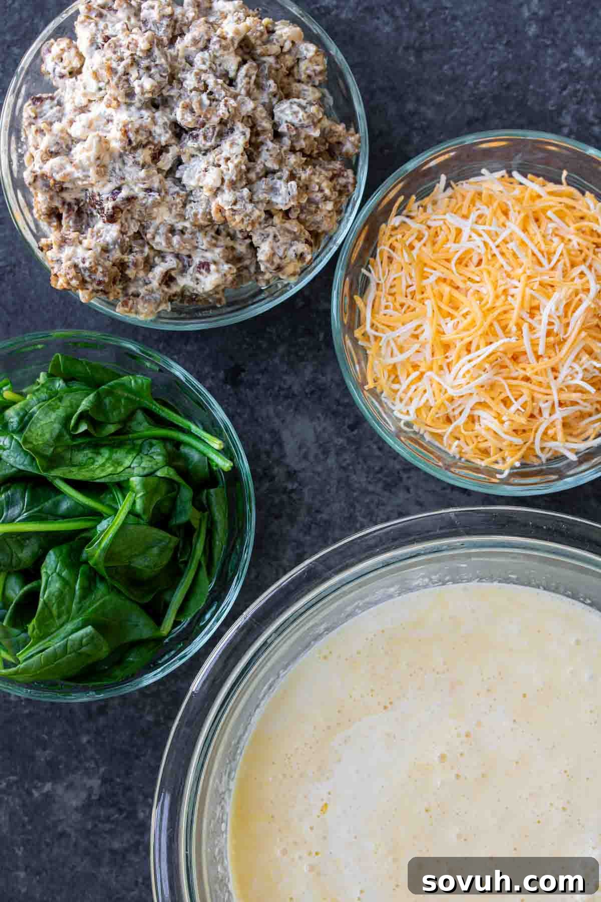 Ingredients for Spinach and Sausage Quiche arranged on a countertop: a bowl of seasoned ground breakfast sausage, a bowl of shredded cheddar cheese, fresh baby spinach leaves, and several large eggs.