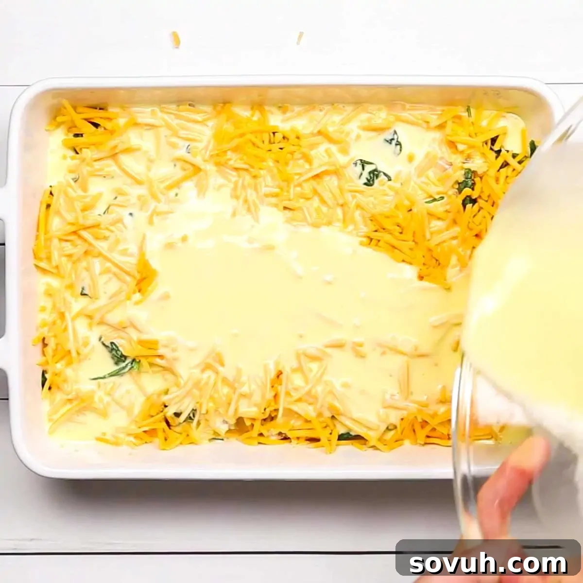 Fresh baby spinach and shredded cheddar cheese layered on top of the sausage mixture in a white baking dish, ready for the egg mixture.
