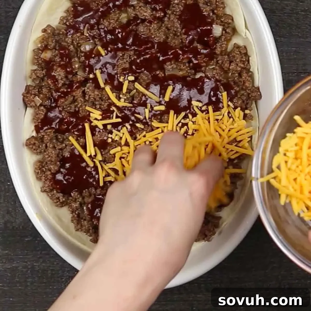 A hand sprinkling shredded cheddar cheese over a layer of beef and enchilada sauce in a casserole dish.